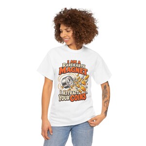 May include: A white short-sleeve t-shirt featuring a graphic of a cartoon magnet with a face surrounded by flying silver and gold coins. The text reads I AM A RARE EARTH MAGNET I ATTRACT YOUR COINS in bold orange and black lettering.