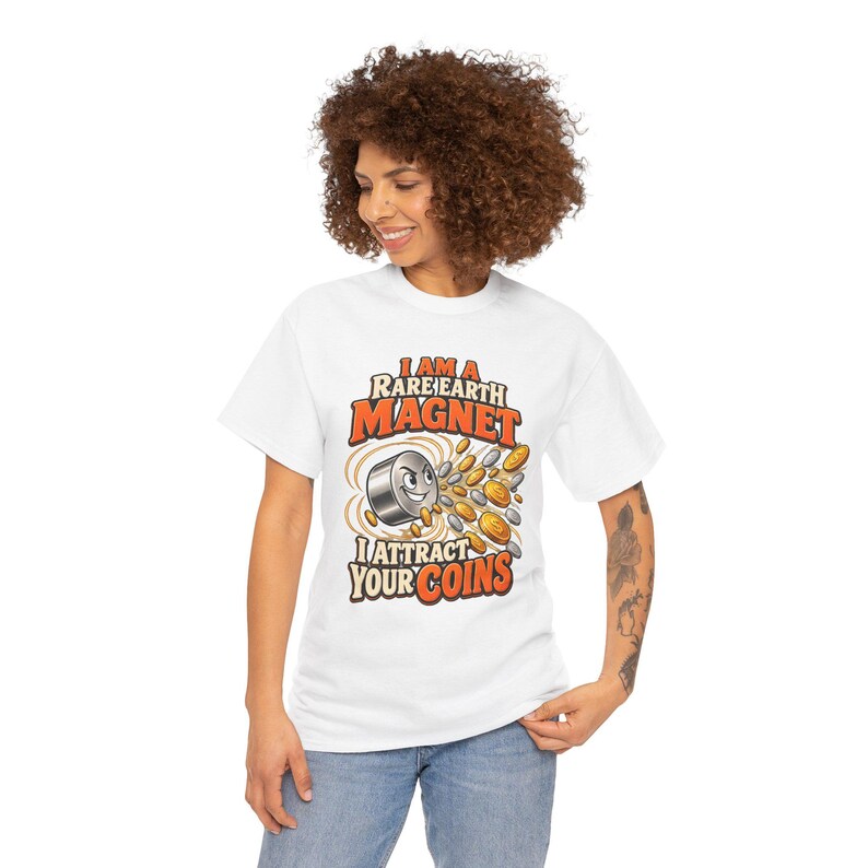 May include: A white short-sleeve t-shirt featuring a graphic of a cartoon magnet with a face surrounded by flying silver and gold coins. The text reads I AM A RARE EARTH MAGNET I ATTRACT YOUR COINS in bold orange and black lettering.