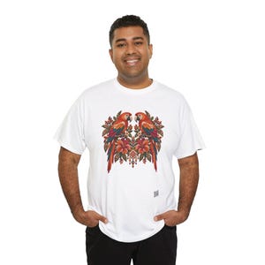 May include: A white short-sleeved t-shirt featuring a central graphic of two symmetrical parrots perched among red hibiscus flowers and green foliage. The design includes intricate patterns in shades of red, orange, blue, and brown.