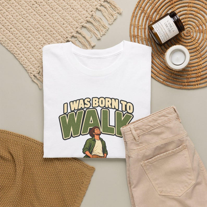 Op de afbeelding: Een wit t-shirt met de tekst I WAS BORN TO WALK in beige en groene blokletters boven een illustratie van een persoon met krullend haar in een groen overhemd en wit ondershirt, die ontspannen omhoog kijkt.