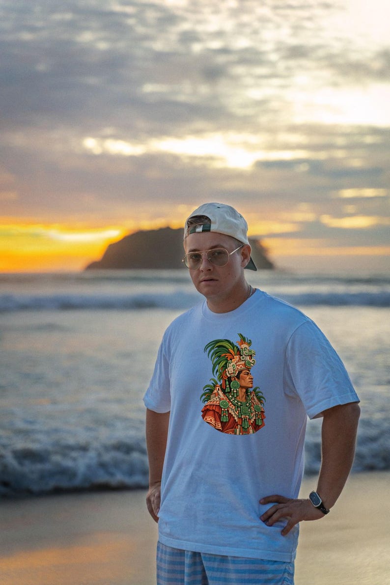 Puede incluir: Una camiseta blanca de manga corta con un estampado gr&aacute;fico colorido de una figura que lleva un elaborado tocado de plumas y joyas tradicionales. La camiseta se lleva con una gorra de b&eacute;isbol y gafas de montura met&aacute;lica.