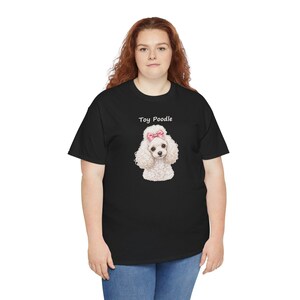 May include: A black short-sleeved t-shirt featuring a graphic of a white toy poodle with a pink bow on its head. The text "Toy Poodle" is printed in white above the dog illustration. The shirt is worn with blue denim jeans.