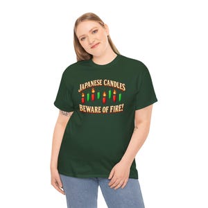 May include: A dark green short-sleeve t-shirt featuring a graphic of red and green candles with flames. The text reads "JAPANESE CANDLES" above the candles and "BEWARE OF FIRE!" below them in a bold, light-coloured, serif font.
