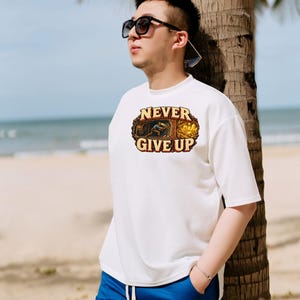 Puede incluir: Una camiseta blanca de manga corta con un estampado gr&aacute;fico de un minero trabajando en una cueva y el texto NEVER GIVE UP en letras may&uacute;sculas de color marr&oacute;n. El dise&ntilde;o est&aacute; enmarcado por un borde ovalado y texturizado.