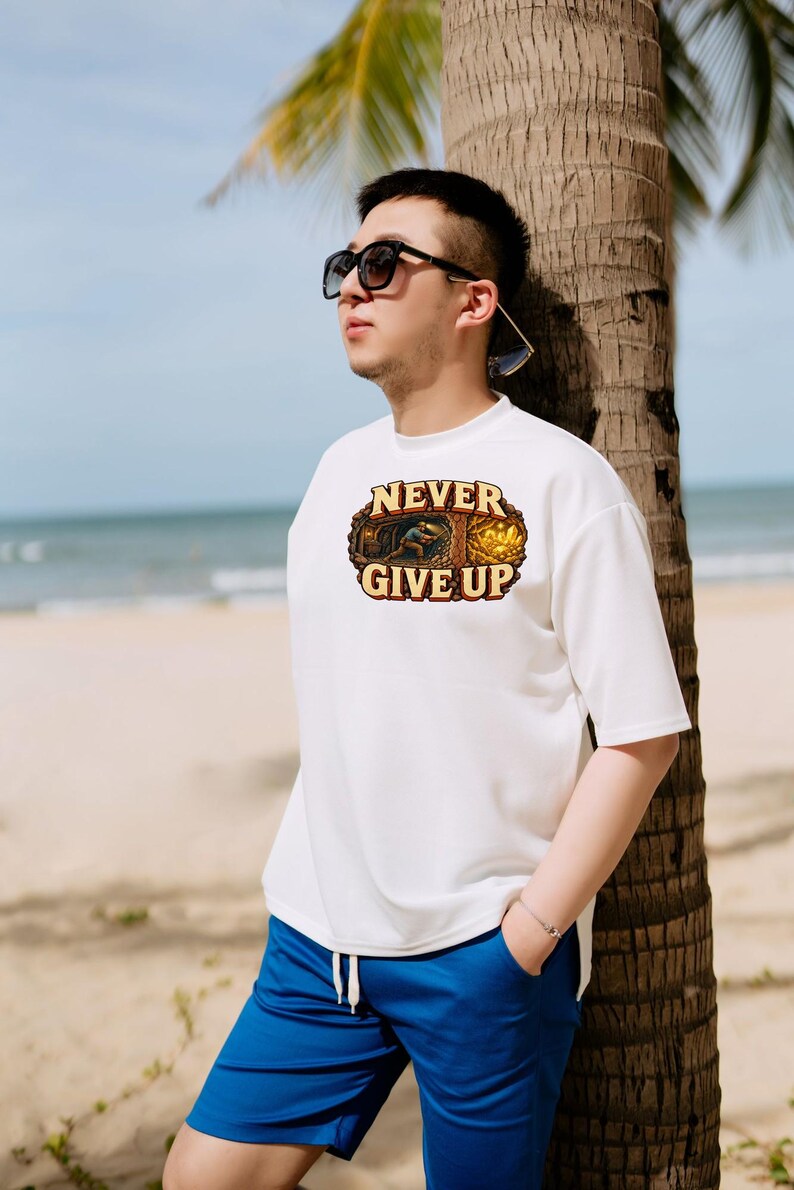 Puede incluir: Una camiseta blanca de manga corta con un estampado gr&aacute;fico de un minero trabajando en una cueva y el texto NEVER GIVE UP en letras may&uacute;sculas de color marr&oacute;n. El dise&ntilde;o est&aacute; enmarcado por un borde ovalado y texturizado.