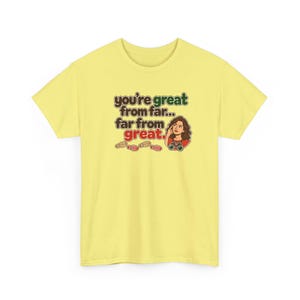 May include: A pale yellow t-shirt featuring a graphic illustration of a person looking through binoculars with the text "you're great from far... far from great." and a series of small footprints printed in brown, green, and red.