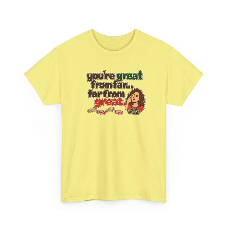 May include: A pale yellow t-shirt featuring a graphic illustration of a person looking through binoculars with the text "you're great from far... far from great." and a series of small footprints printed in brown, green, and red.