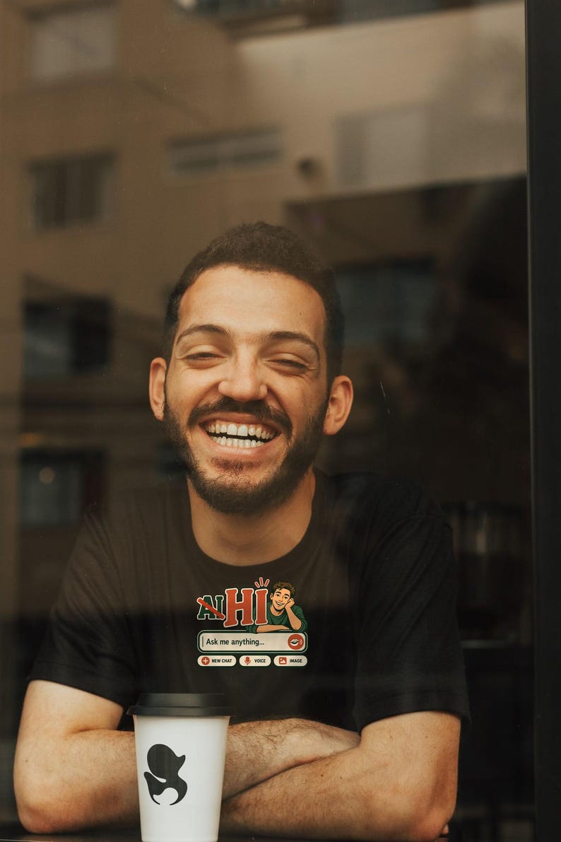Puede incluir: Una camiseta negra con un estampado gr&aacute;fico que incluye el texto HI, la ilustraci&oacute;n de una persona y un cuadro de interfaz de chat con el texto Ask me anything, junto con iconos de chat, voz e imagen, acompa&ntilde;ada de una taza blanca.