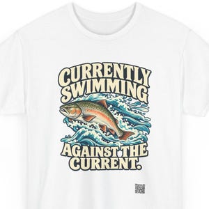 May include: A white t-shirt featuring a graphic of a trout jumping through blue water waves. The text above the fish reads CURRENTLY SWIMMING and the text below reads AGAINST THE CURRENT. A small QR code is printed near the bottom right corner.