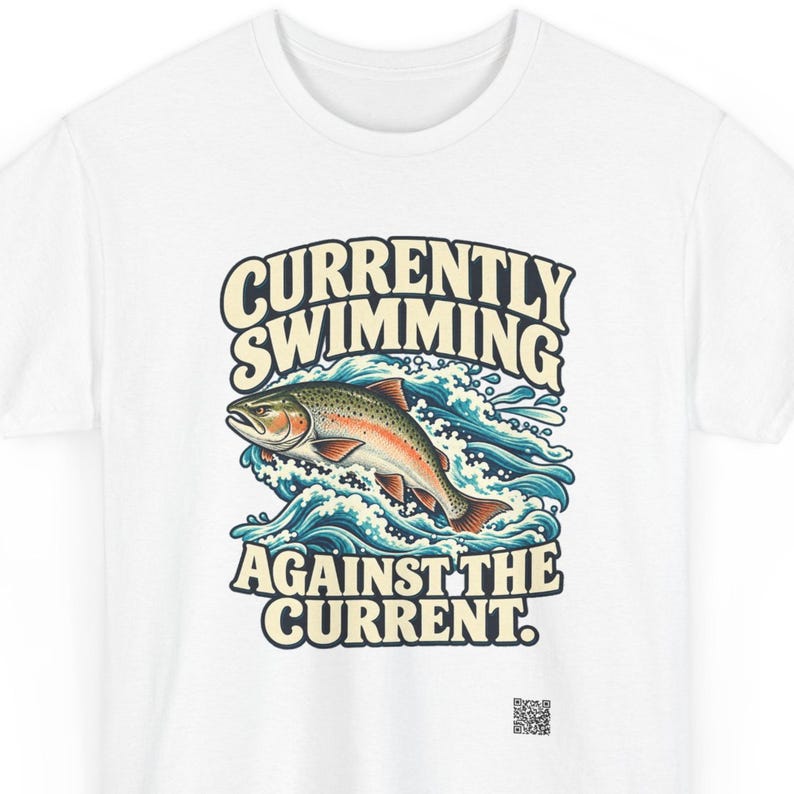 May include: A white t-shirt featuring a graphic of a trout jumping through blue water waves. The text above the fish reads CURRENTLY SWIMMING and the text below reads AGAINST THE CURRENT. A small QR code is printed near the bottom right corner.