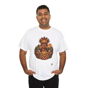 May include: A white short-sleeved t-shirt featuring a central graphic illustration of a person wearing an elaborate feathered headdress and gold jewelry, set against a backdrop of mountains and a sun, with a small square logo at the bottom.