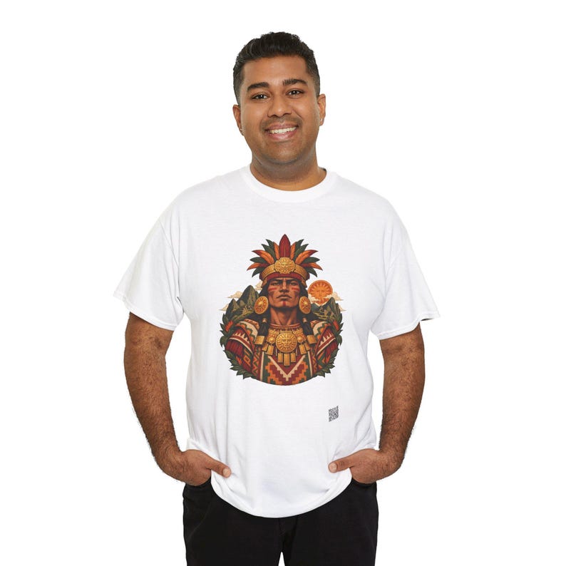 May include: A white short-sleeved t-shirt featuring a central graphic illustration of a person wearing an elaborate feathered headdress and gold jewelry, set against a backdrop of mountains and a sun, with a small square logo at the bottom.