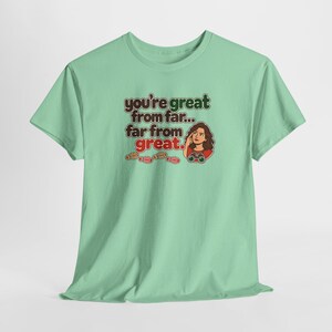 May include: A light green short-sleeve t-shirt featuring a graphic of a person looking through binoculars with the text "you're great from far... far from great." and four small red and brown shoe print icons below the text.