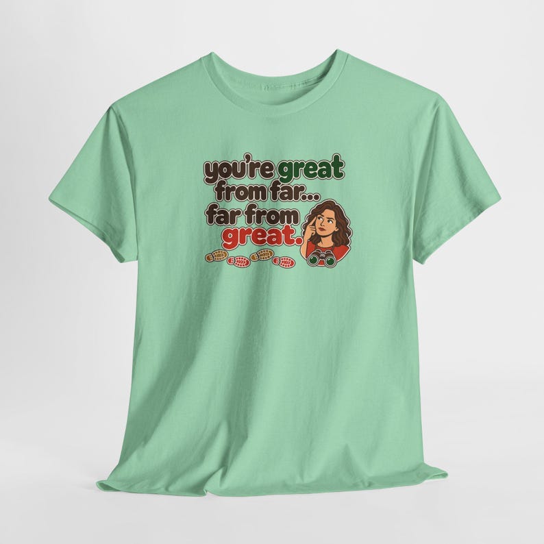 May include: A light green short-sleeve t-shirt featuring a graphic of a person looking through binoculars with the text "you're great from far... far from great." and four small red and brown shoe print icons below the text.