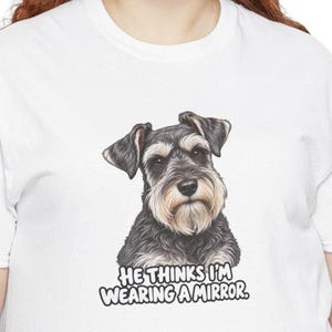 May include: A white t-shirt featuring a detailed illustration of a gray and black Schnauzer dog. Below the dog, the text reads: HE THINKS I'M WEARING A MIRROR. The graphic is centered on the chest of the shirt, showcasing the dog's face.