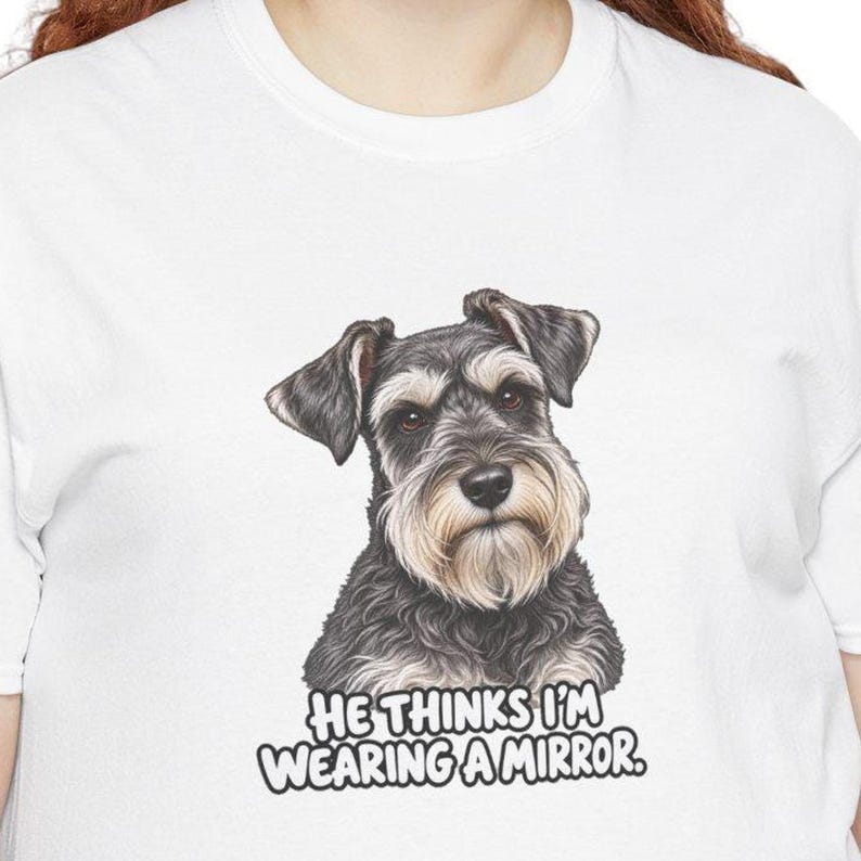 May include: A white t-shirt featuring a detailed illustration of a gray and black Schnauzer dog. Below the dog, the text reads: HE THINKS I'M WEARING A MIRROR. The graphic is centered on the chest of the shirt, showcasing the dog's face.