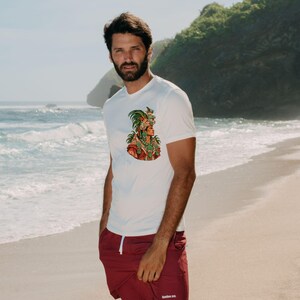 Puede incluir: Una camiseta blanca de manga corta con un gr&aacute;fico colorido de un guerrero azteca con un tocado de plumas en el pecho, combinada con pantalones cortos de color granate liso con un peque&ntilde;o logotipo blanco en la pierna derecha.