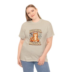 May include: A beige short-sleeve t-shirt featuring a graphic of an orange tabby cat sitting in a lotus position. The text above and below the cat reads, "LESS DRAMA, MORE KARMA." The design includes small star and swirl accents.