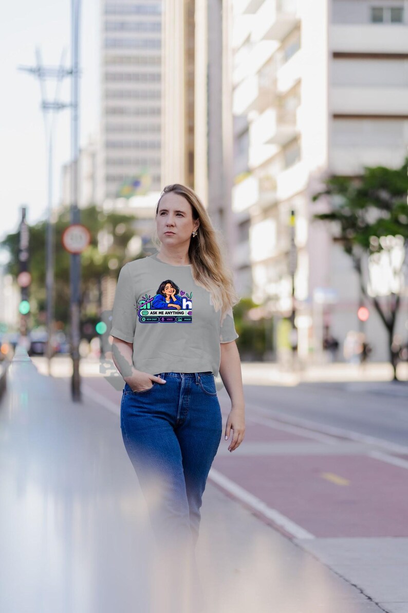 May include: A light gray short-sleeved t-shirt featuring a graphic print of a person in a blue shirt with the text "ASK ME ANYTHING" and various icons. The shirt has a crew neckline and a boxy, cropped fit, worn with dark blue denim jeans.