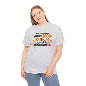May include: A light gray short-sleeve t-shirt featuring the text "from 100 feet away, you're 100%." in black and orange, with three shoe print graphics in orange and red, followed by "sadly, every foot closer knocks off 1%." in black and red.