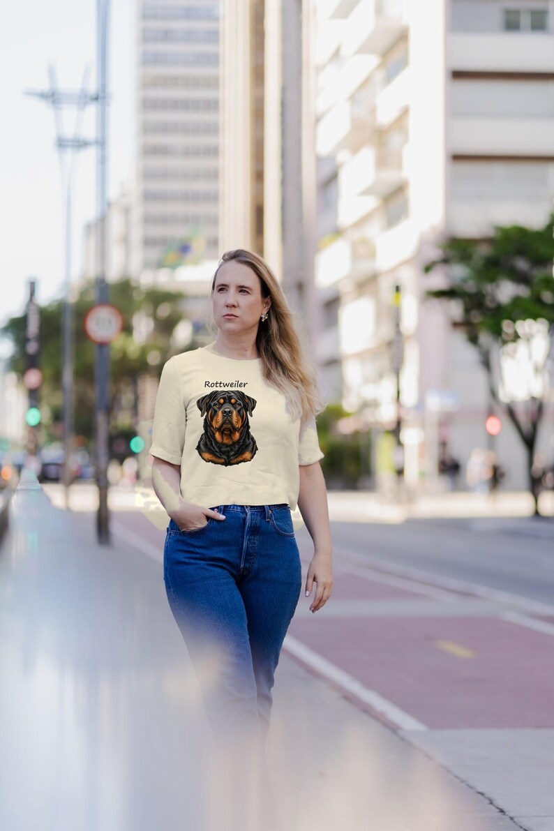 May include: A cream-colored short-sleeve t-shirt featuring a graphic illustration of a Rottweiler dog head centered on the chest with the word Rottweiler printed in black text above the dog's head.