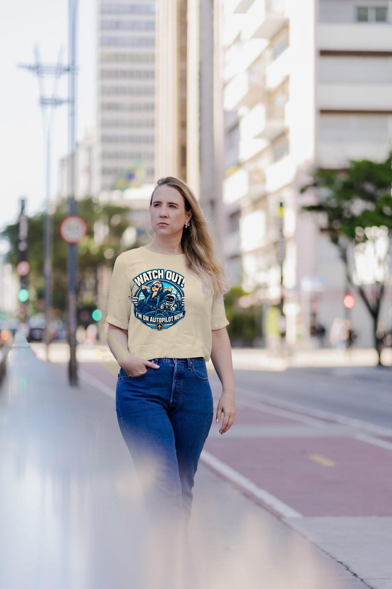 May include: A beige short-sleeved t-shirt featuring a circular graphic print of a pilot wearing sunglasses and a small robot. The text reads WATCH OUT! I'M ON AUTOPILOT NOW. The shirt is paired with blue denim jeans.