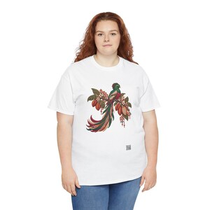 May include: A white short-sleeved t-shirt featuring a central graphic of a quetzal bird perched on a branch with cacao pods. The bird has vibrant green, red, and white plumage with a long tail. Geometric patterns and small flowers surround the bird.