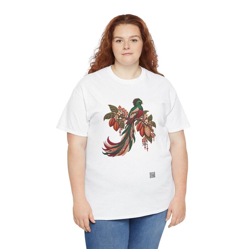 May include: A white short-sleeved t-shirt featuring a central graphic of a quetzal bird perched on a branch with cacao pods. The bird has vibrant green, red, and white plumage with a long tail. Geometric patterns and small flowers surround the bird.