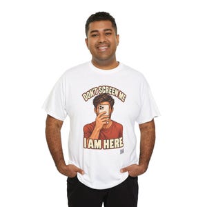 Op de afbeelding: Een wit T-shirt met korte mouwen en een afbeelding van een persoon die een smartphone voor het gezicht houdt. De tekst boven en onder de afbeelding luidt "DON'T SCREEN ME I AM HERE" in dikke, bruine blokletters.