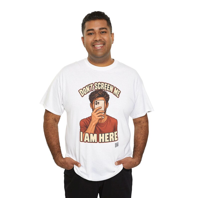 Op de afbeelding: Een wit T-shirt met korte mouwen en een afbeelding van een persoon die een smartphone voor het gezicht houdt. De tekst boven en onder de afbeelding luidt "DON'T SCREEN ME I AM HERE" in dikke, bruine blokletters.