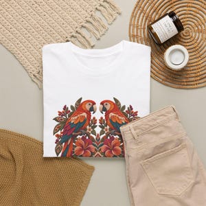 May include: A white t-shirt featuring a symmetrical graphic print of two colorful parrots perched among tropical flowers and foliage. The parrots have red, orange, and blue feathers, and the floral design includes large orange hibiscus blossoms and small berries.