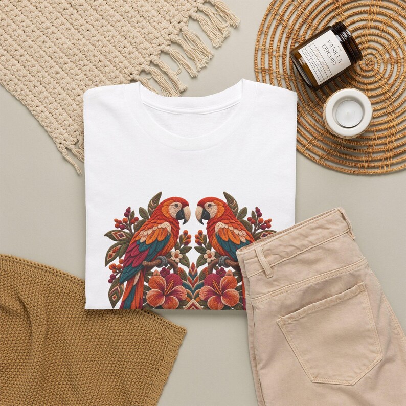May include: A white t-shirt featuring a symmetrical graphic print of two colorful parrots perched among tropical flowers and foliage. The parrots have red, orange, and blue feathers, and the floral design includes large orange hibiscus blossoms and small berries.