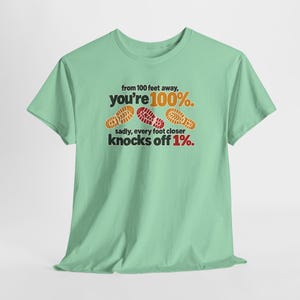 May include: A light green short-sleeve t-shirt featuring the text from 100 feet away, you're 100%. sadly, every foot closer knocks off 1%. The design includes three colorful shoe sole prints in orange, red, and yellow across the chest area.