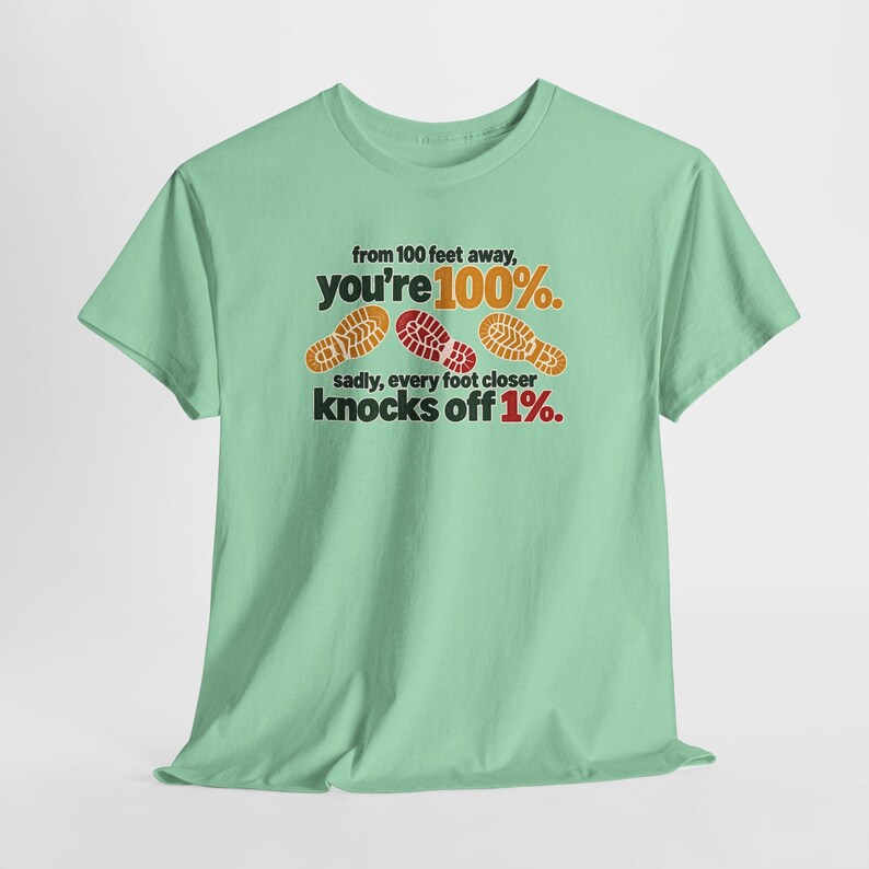 May include: A light green short-sleeve t-shirt featuring the text from 100 feet away, you're 100%. sadly, every foot closer knocks off 1%. The design includes three colorful shoe sole prints in orange, red, and yellow across the chest area.