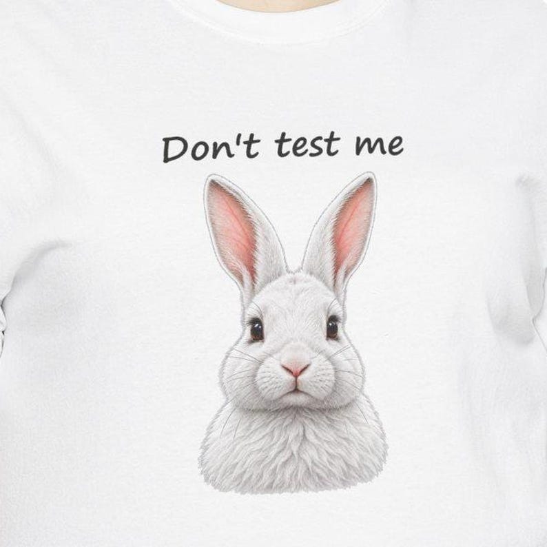 May include: A white t-shirt featuring a graphic of a fluffy white rabbit with pink-tinted ears. Above the rabbit, the text "Don't test me" is printed in a simple black sans-serif font. The design is centered on the front of the garment.