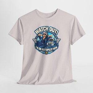 May include: A light gray t-shirt featuring a circular graphic of a pilot wearing sunglasses and headphones sitting next to a small robot in a cockpit. The text reads WATCH OUT! I'M ON AUTOPILOT NOW. The design includes blue and white accents.