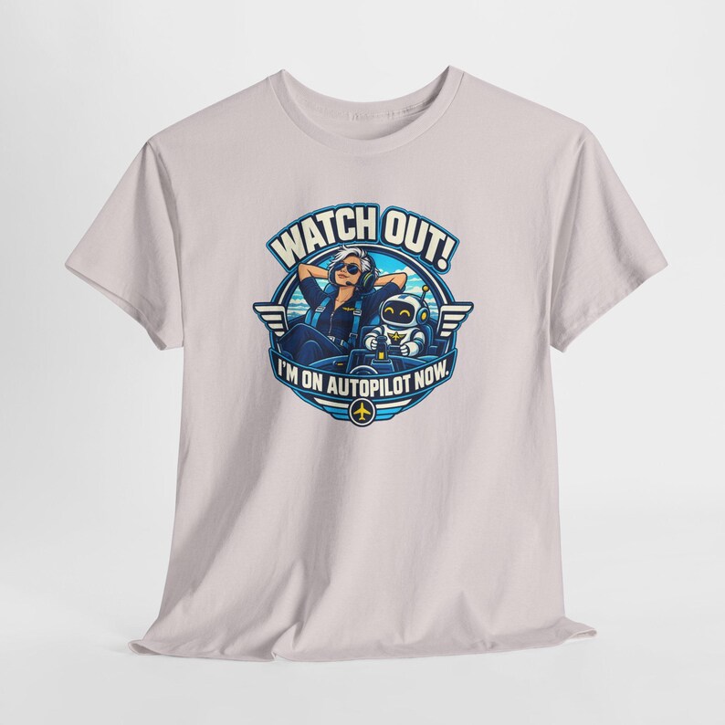 May include: A light gray t-shirt featuring a circular graphic of a pilot wearing sunglasses and headphones sitting next to a small robot in a cockpit. The text reads WATCH OUT! I'M ON AUTOPILOT NOW. The design includes blue and white accents.