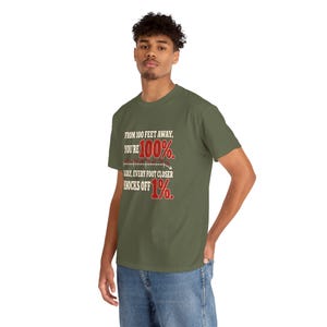 May include: An olive green short-sleeve t-shirt featuring white and red text that reads: FROM 100 FEET AWAY, YOU'RE 100%. SADLY, EVERY FOOT CLOSER KNOCKS OFF 1%. The design includes a small graphic of a downward-sloping line graph.