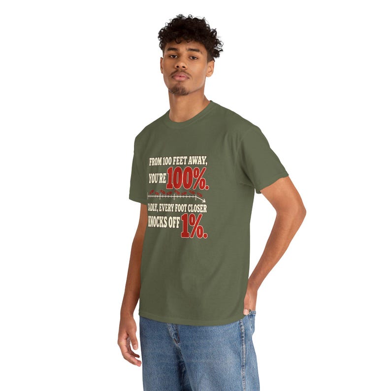 May include: An olive green short-sleeve t-shirt featuring white and red text that reads: FROM 100 FEET AWAY, YOU'RE 100%. SADLY, EVERY FOOT CLOSER KNOCKS OFF 1%. The design includes a small graphic of a downward-sloping line graph.