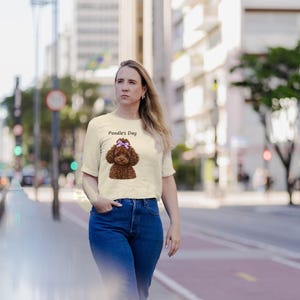 May include: A cream-colored short-sleeve t-shirt featuring a graphic of a brown poodle with a purple bow on its head, accompanied by the text "Poodle's Day" printed in black above the dog illustration.
