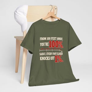 May include: An olive green t-shirt featuring the text: FROM 100 FEET AWAY, YOU'RE 100%. SADLY, EVERY FOOT CLOSER KNOCKS OFF 1%. The design includes a graphic of a downward-sloping line with red numbers and small red oval shapes.