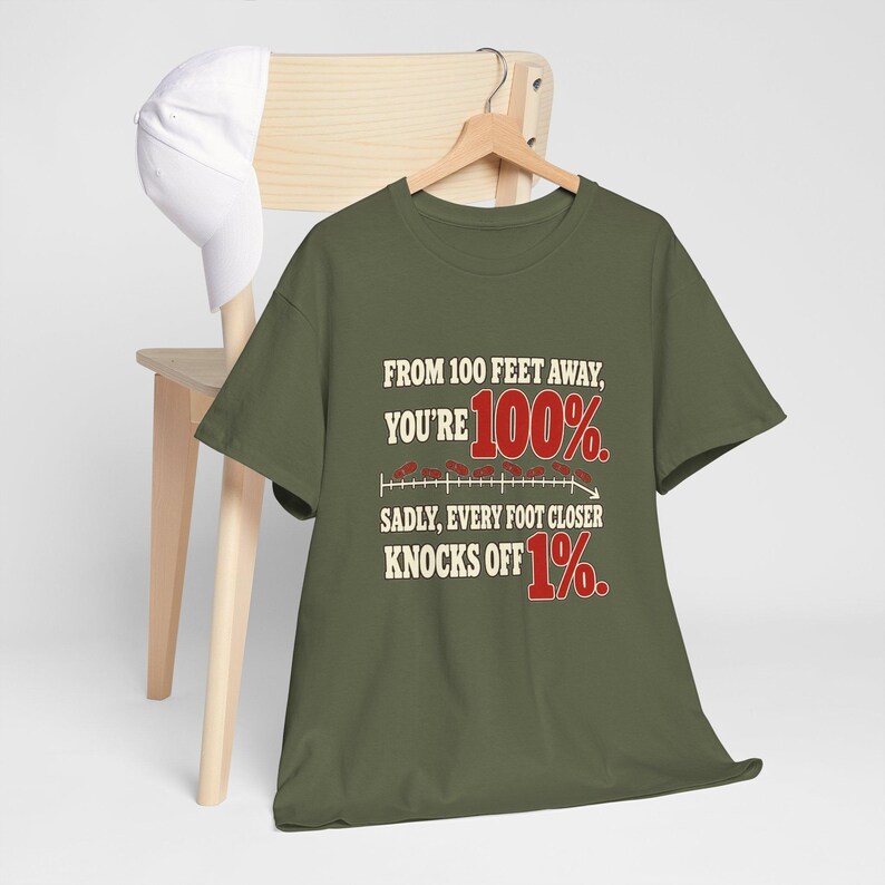 May include: An olive green t-shirt featuring the text: FROM 100 FEET AWAY, YOU'RE 100%. SADLY, EVERY FOOT CLOSER KNOCKS OFF 1%. The design includes a graphic of a downward-sloping line with red numbers and small red oval shapes.