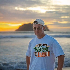 May include: A white short-sleeved t-shirt featuring the text "YOU ARE WELCOME... TO LEAVE!" printed in green and orange lettering with a small graphic of an open door next to the word LEAVE on the front chest area.