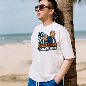 May include: A white short-sleeved t-shirt featuring a graphic illustration of a person with a hand raised in a stop gesture, a cigarette, and the text "SMOKE THERE I BREATHE HERE" in orange and blue, paired with blue drawstring shorts.
