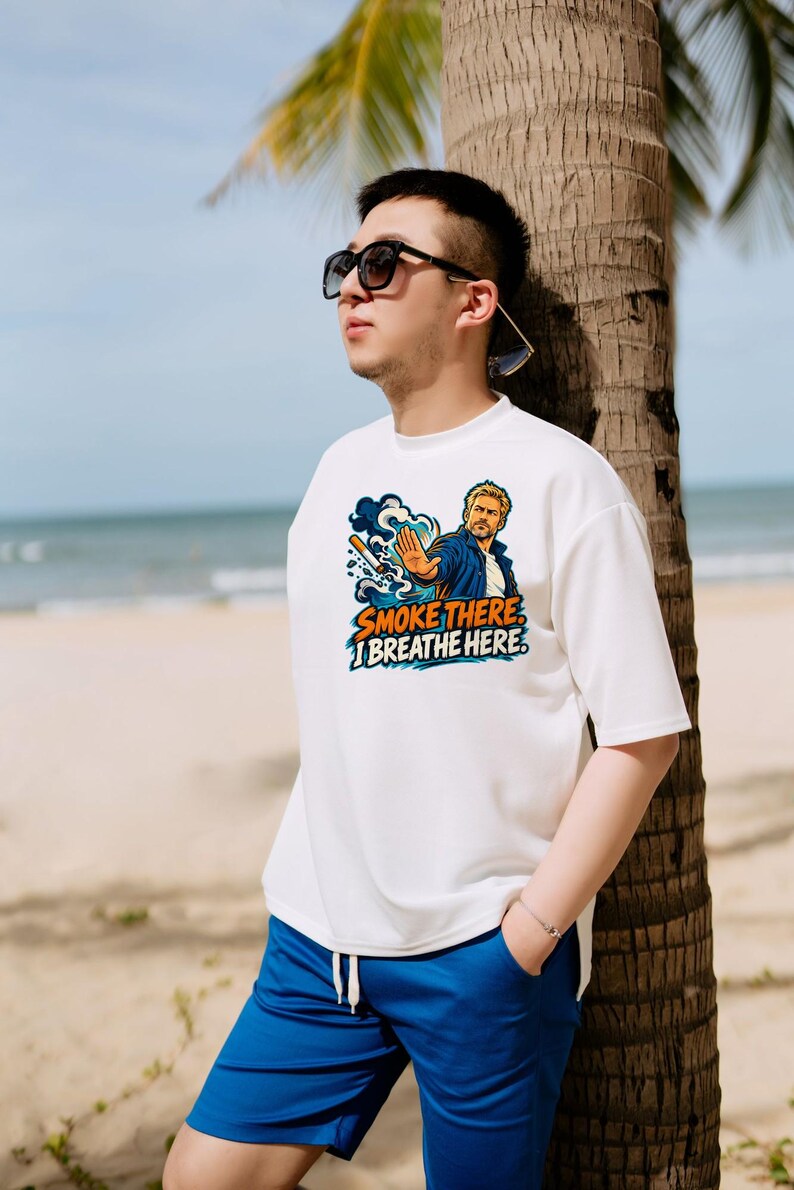 May include: A white short-sleeved t-shirt featuring a graphic illustration of a person with a hand raised in a stop gesture, a cigarette, and the text "SMOKE THERE I BREATHE HERE" in orange and blue, paired with blue drawstring shorts.