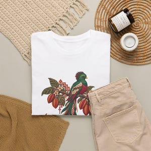May include: A white t-shirt featuring a graphic illustration of a green and red quetzal bird perched on a branch with cocoa pods and white flowers. The design includes geometric patterns in red and brown tones near the bird.