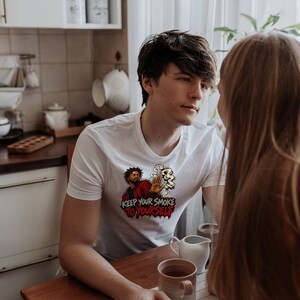 May include: A white short-sleeved t-shirt featuring a graphic illustration of a person in a red jacket holding up a hand with smoke swirling around it. Below the illustration, the text reads KEEP YOUR SMOKE TO YOURSELF in bold letters.