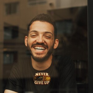 Puede incluir: Una camiseta negra de manga corta con un estampado gr&aacute;fico de una persona cavando en una mina y el texto "NEVER GIVE UP" encima. La camiseta se lleva con un vaso de caf&eacute; desechable blanco con un logotipo negro.