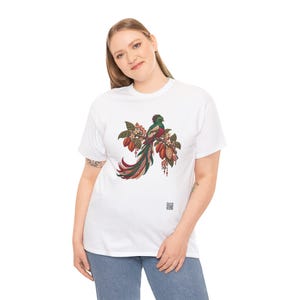 May include: A white short-sleeved t-shirt featuring a central graphic of a quetzal bird perched on a branch with cacao pods and leaves. The bird has green, red, and long tail feathers. A small square QR code is printed near the bottom right.