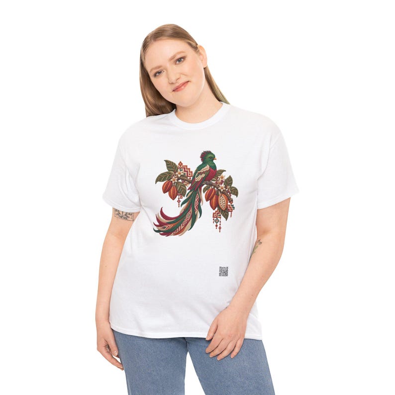 May include: A white short-sleeved t-shirt featuring a central graphic of a quetzal bird perched on a branch with cacao pods and leaves. The bird has green, red, and long tail feathers. A small square QR code is printed near the bottom right.